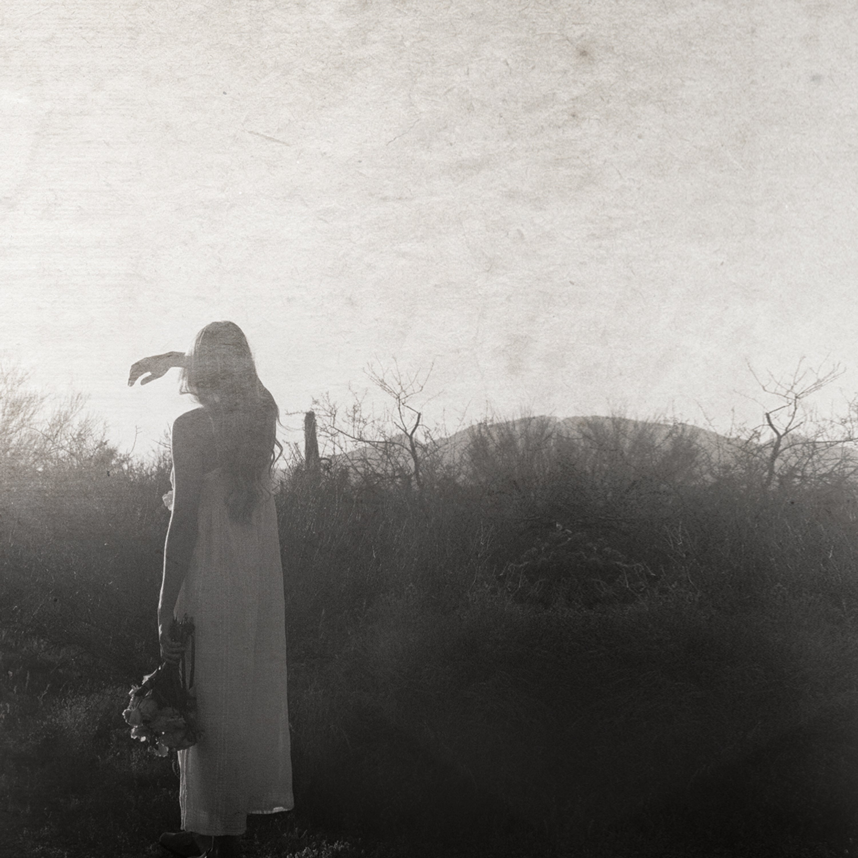 A monochrome, textured photograph of a woman in a long white dress standing outdoors in a field, holding a bouquet and looking toward the bright horizon.