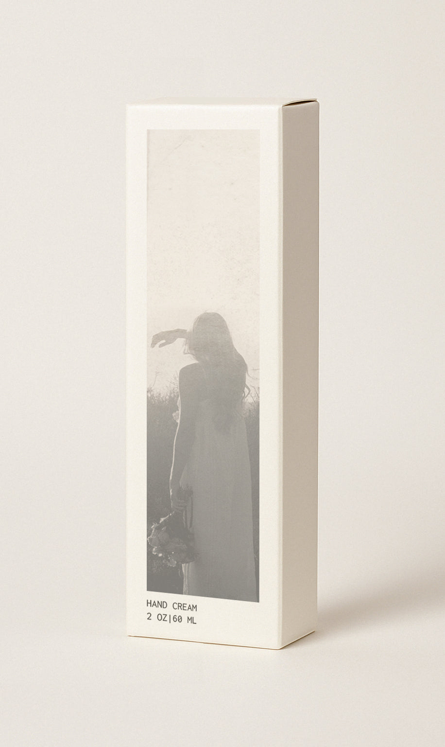 Product packaging image showing the box that the hand cream comes in. Box measures 2" wide x 1.25" deep x 6.5" high. Box is cream colored with sepia toned image of woman in the landscape. Box also has the text 'Hand cream' and '20 oz | 60 ml'. Image has cream background.