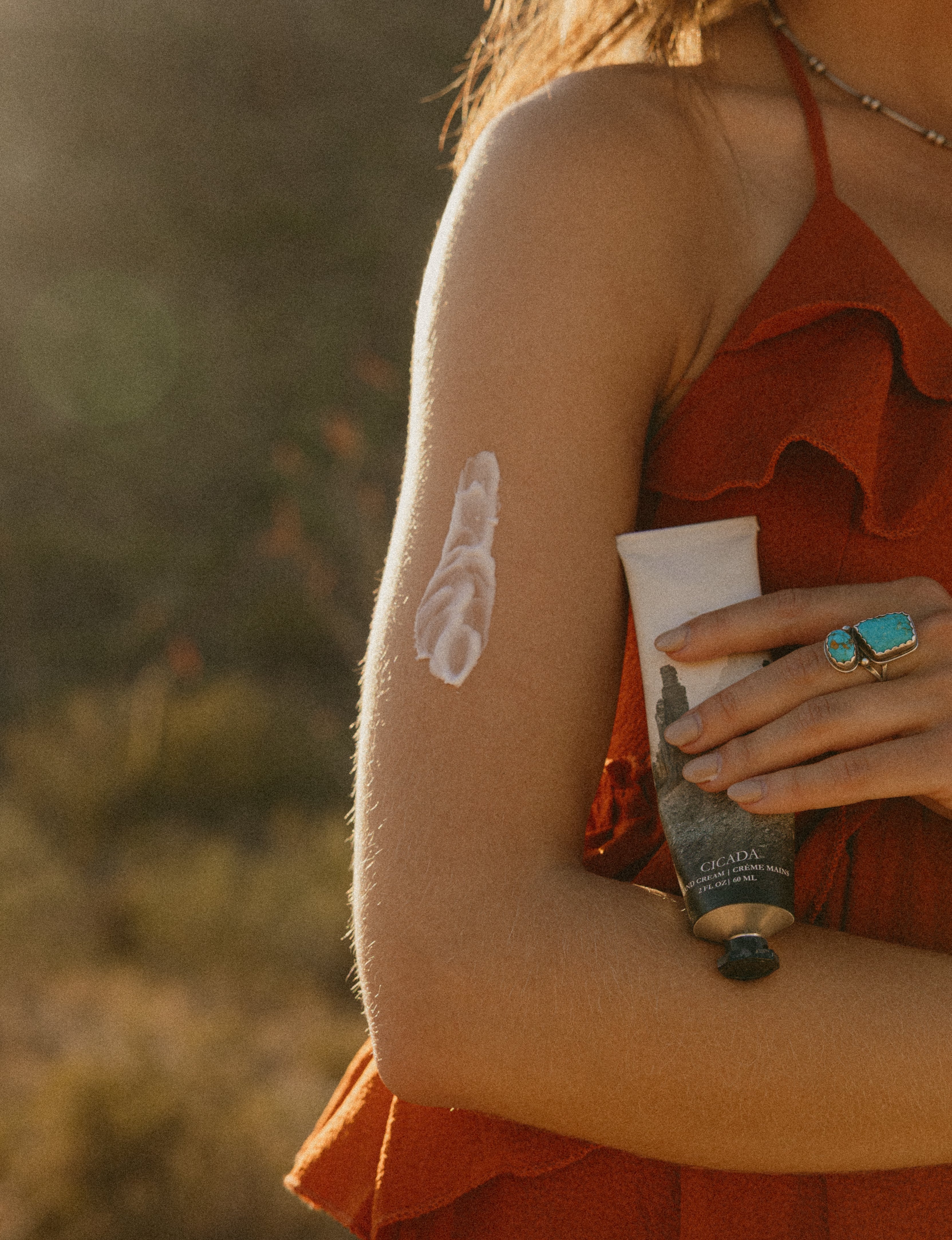 Close-up of a girl in red dress and turquoise ring in the desert and holding a CICADA hand cream tube with lotion applied to their arm.