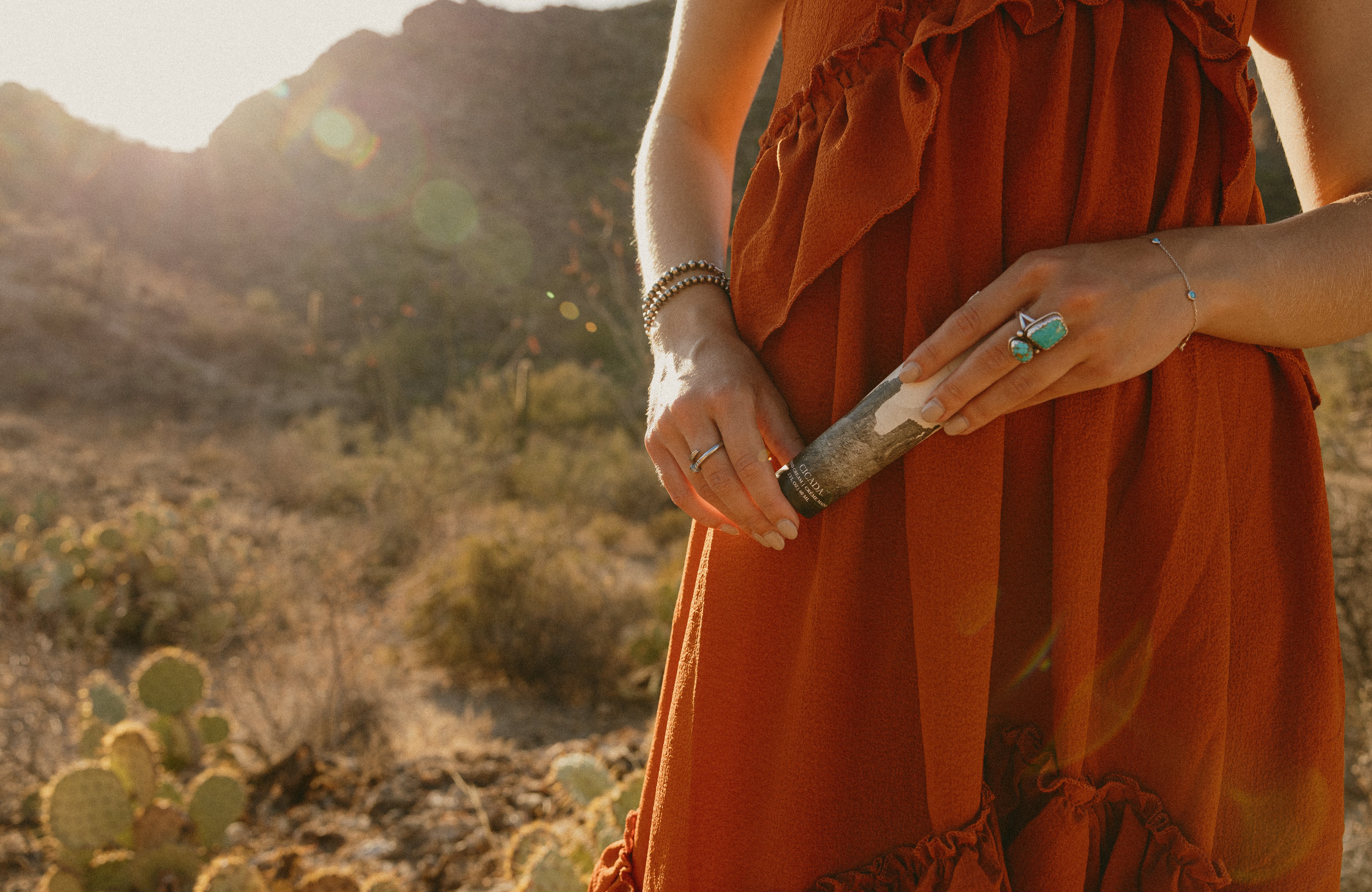 Lifestyle image of woman standing in desert landscape holding CICADA hand cream in her hands. Woman is wearing red dress.