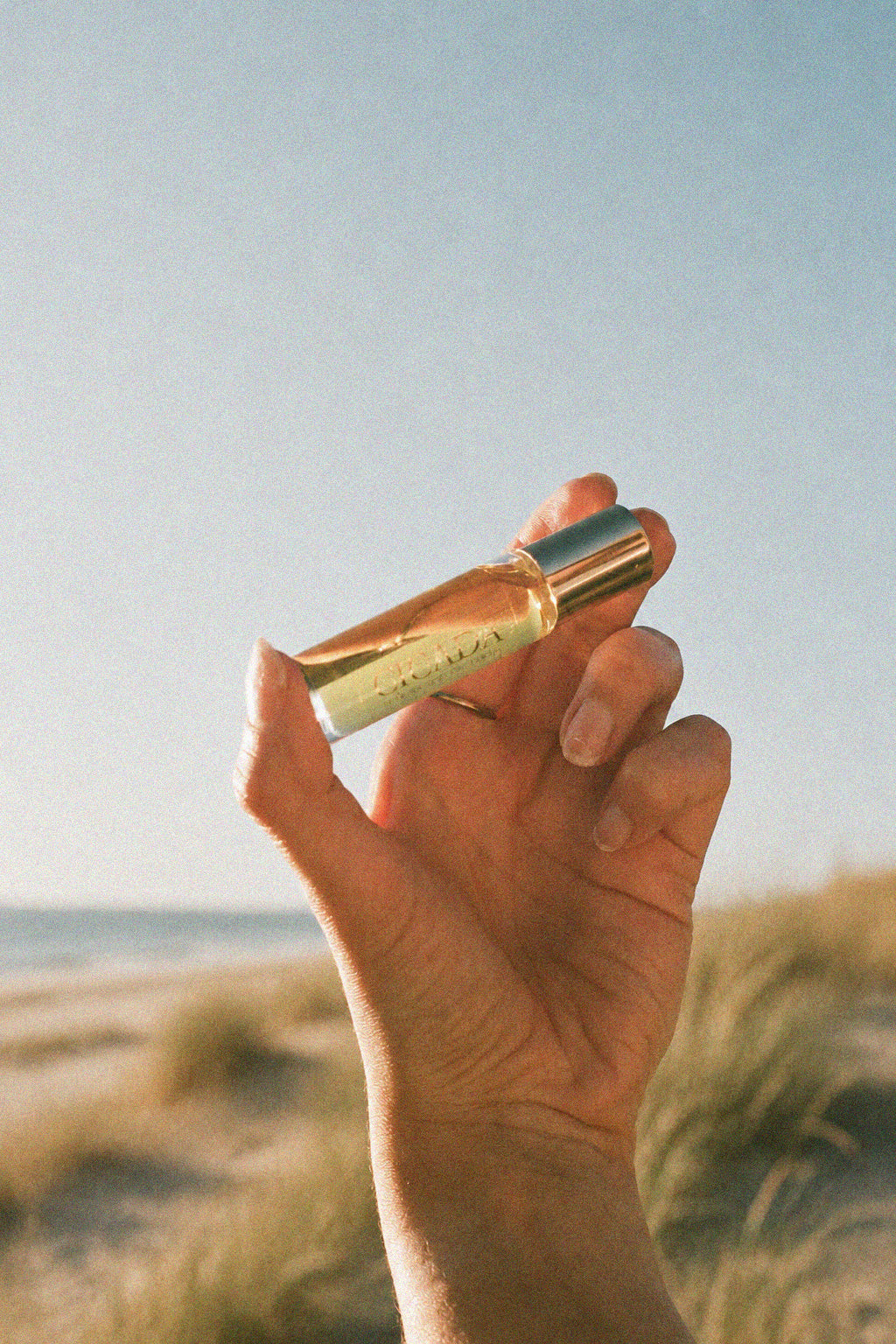 A hand holding a CICADA glass perfume oil bottle with a metallic cap, set against a blurred beach dune background.