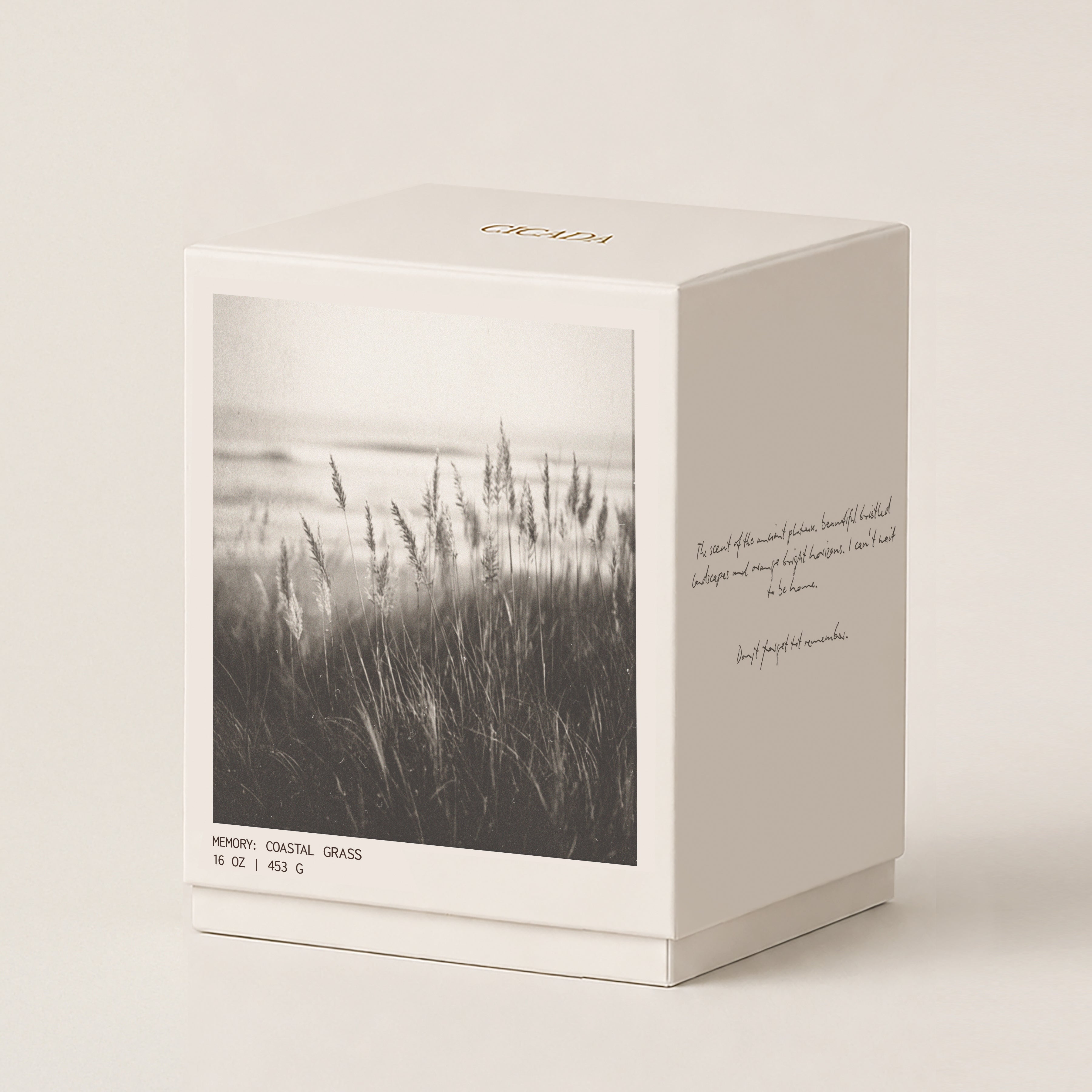 A cream colored candle box with a black-and-white coastal grass photograph on the front, photographed on a clean warm off-white background. The box has small gold lettering near the top and handwritten-style text on the side.