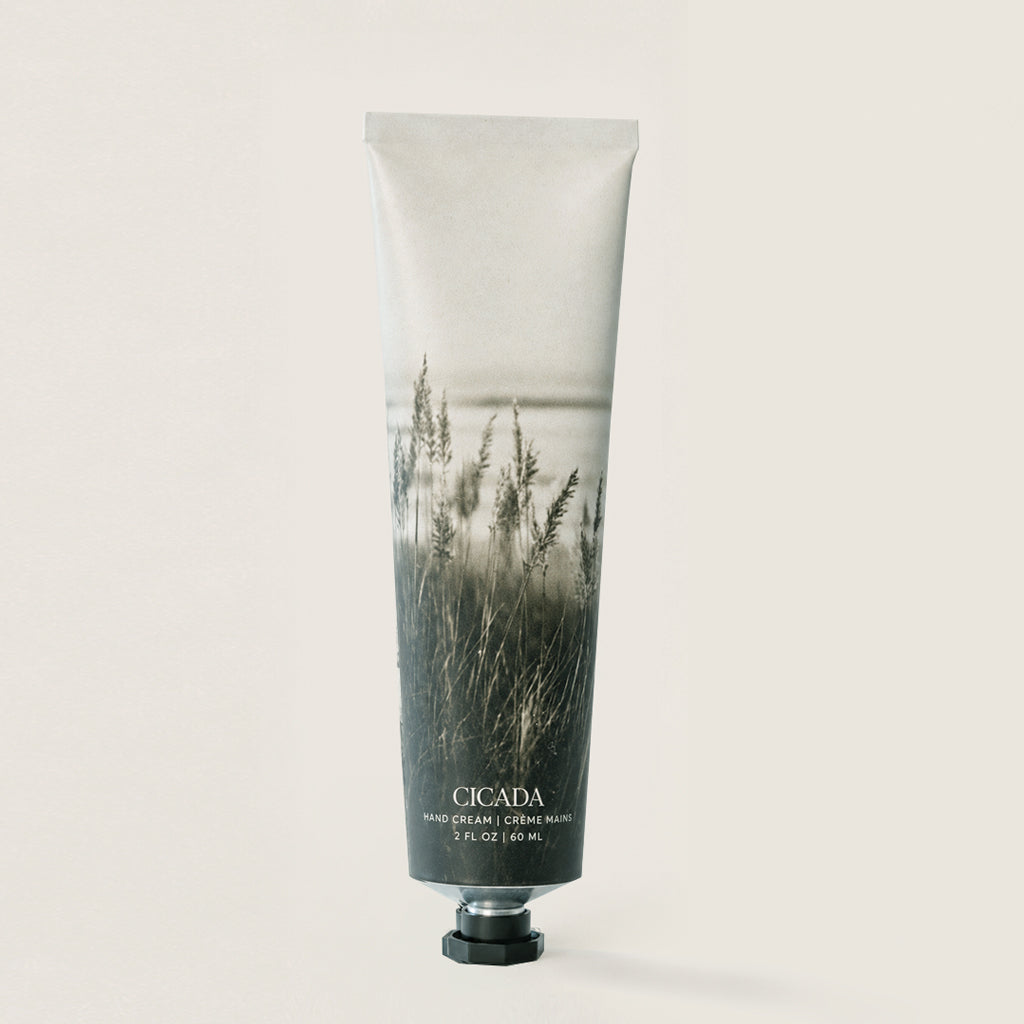 A single tube of Cicada Hand cream on a solid light beige background. The tube features a photographic print of tall grasses in misty water, fading from light gray at the top to dark gray/black at the bottom, with the brand name 'CICADA' printed in white near the cap.