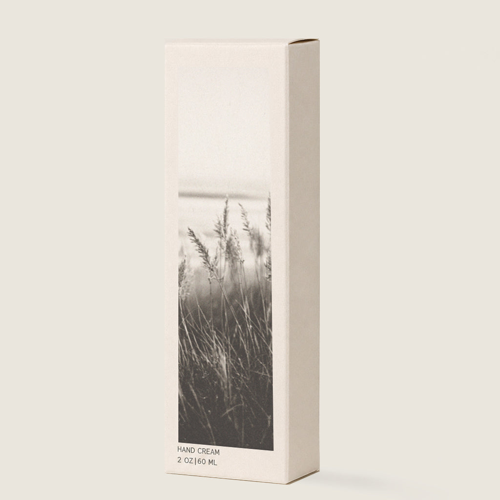 A minimalist rectangular hand cream box with a black and white photograph of beach grass on the front, set against a solid light beige background.