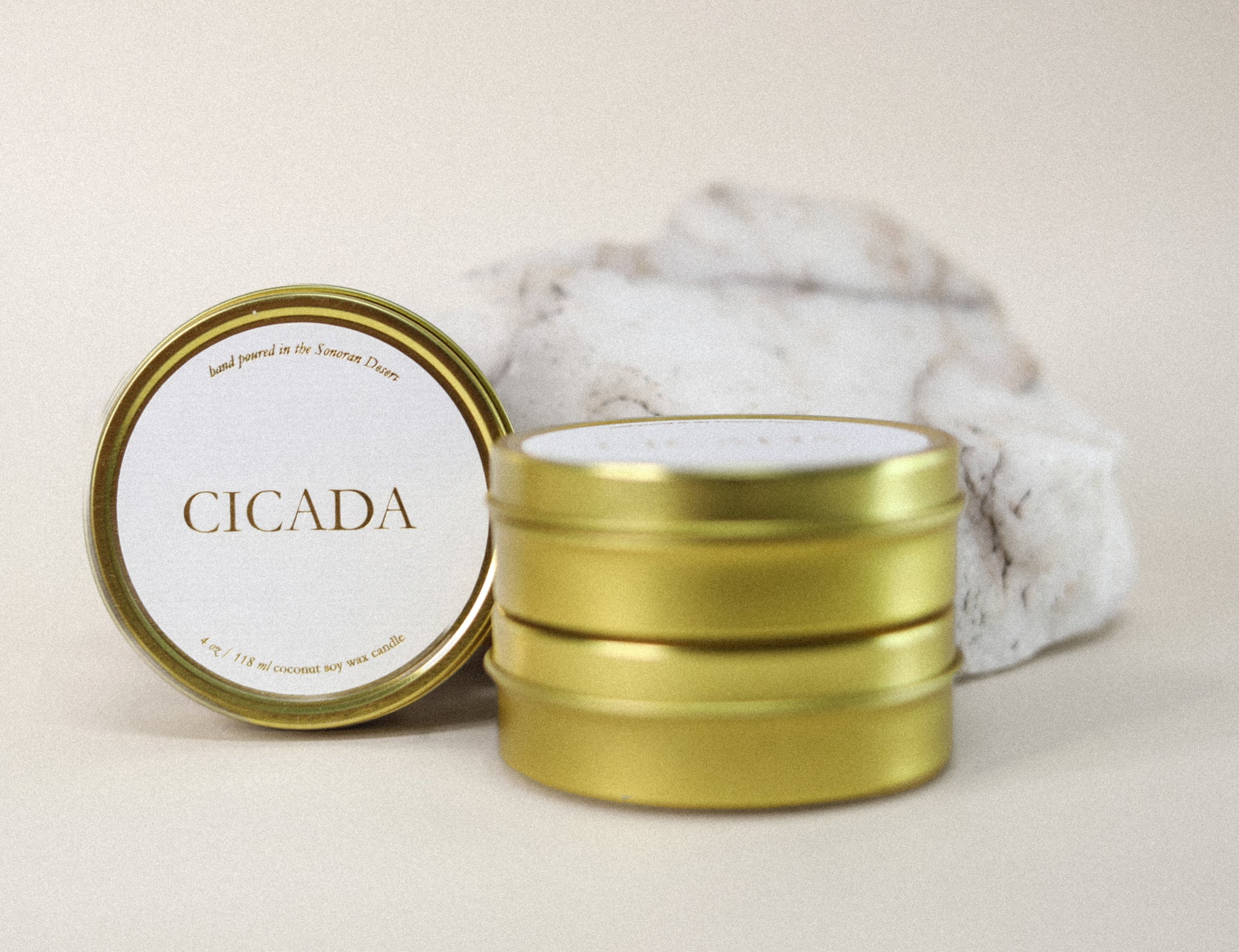 Product image of 4oz gold candles stacked. And one old tin candle turned on side to see CICADA label. Rock and cream background. 