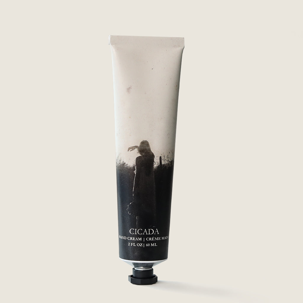 Product image of the CICADA hand cream. Image shows aluminum tube with CICADA logo on it. Hand cream tube has black and white image of woman standing in the landscape. Hand cream tube also shows the text 'HAND CREAM (in English and French) as well as 2oz | 60 ml. Image has cream back ground