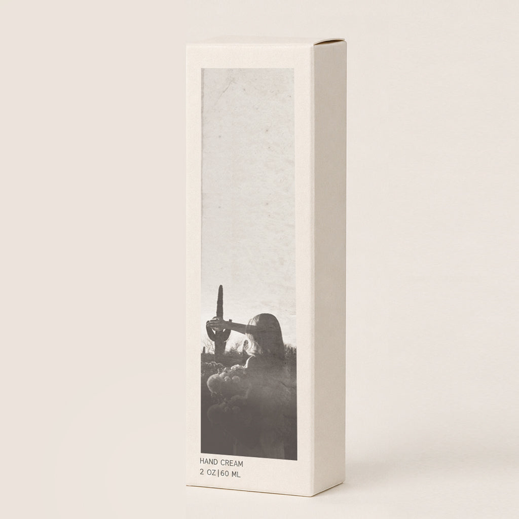 A tall, cream colored hand cream, featuring a vertical black-and-white photographic print on the front of a woman and a cactus. The box is photographed on a clean, solid, warm off-white background.