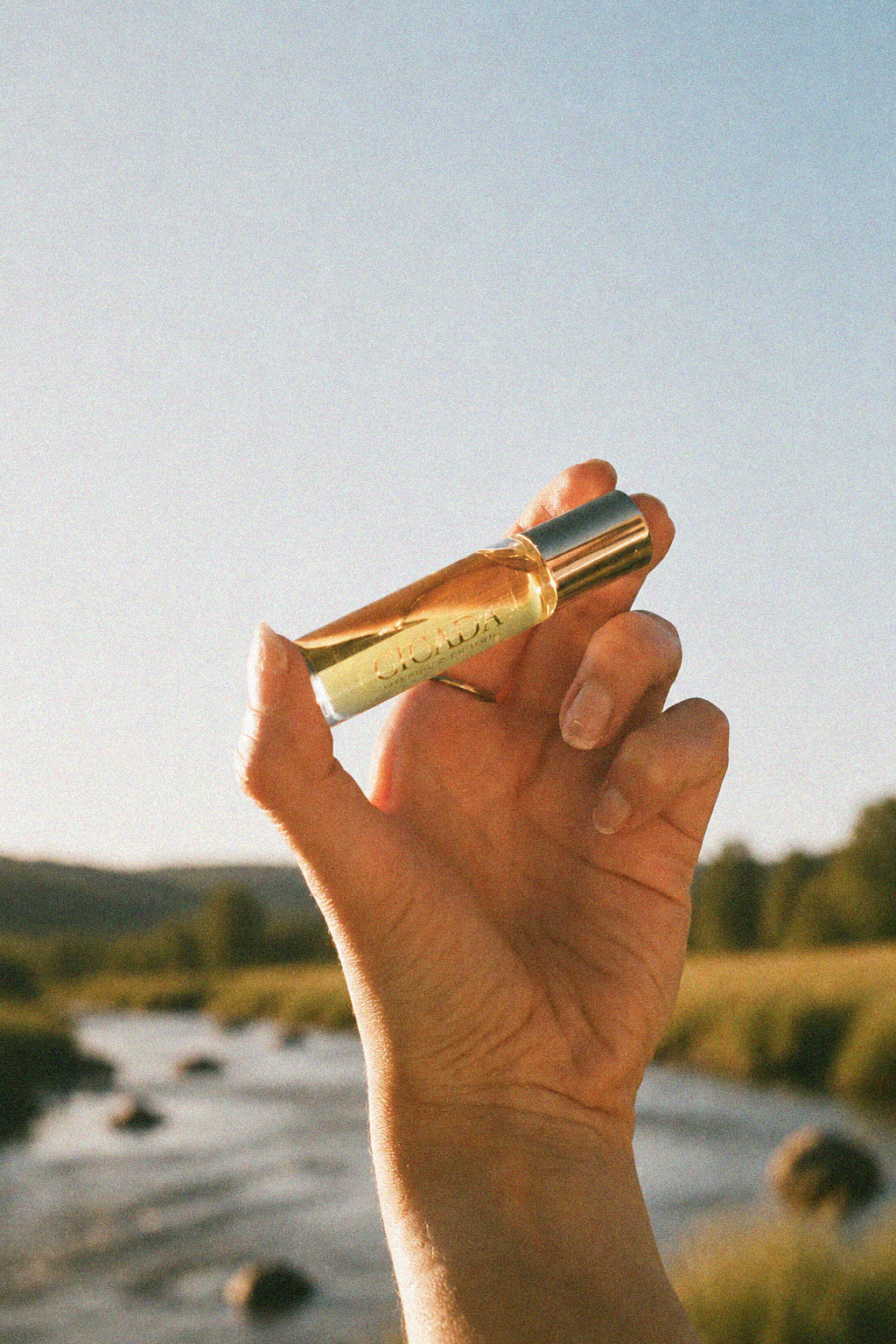 A hand holding a small cylindrical bottle of CICADA perfume oil with a gold cap, set against a blurred outdoor landscape with a river and trees under a clear blue sky.