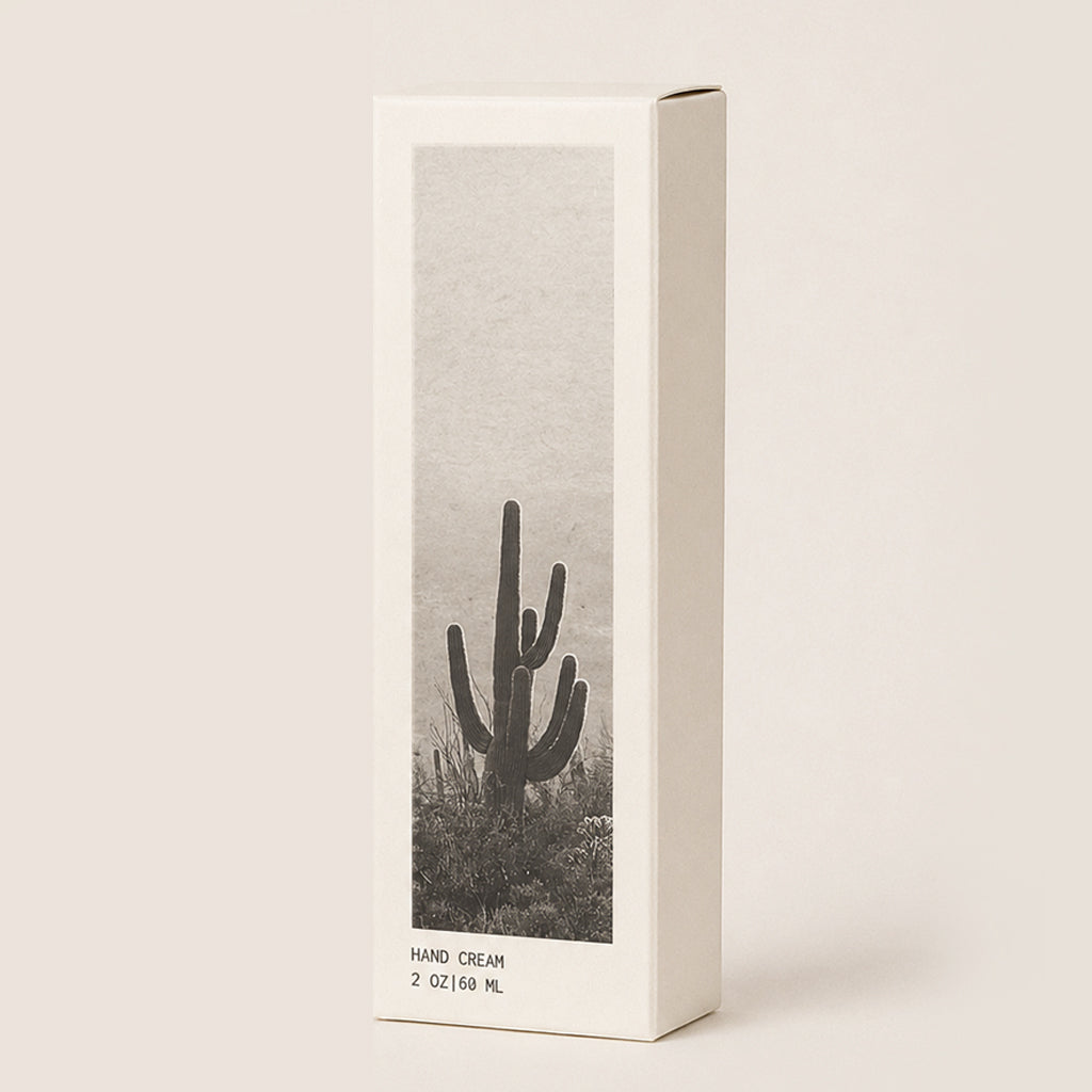A tall rectangular hand cream box with a minimalist design, featuring a black and white desert scene with a saguaro cactus on the front panel, set against a solid light beige background.